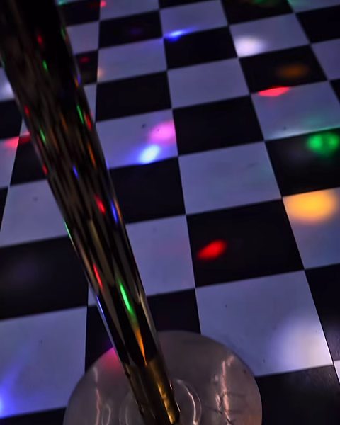 The dance pole on the checkerboard floor, with coloured disco lights