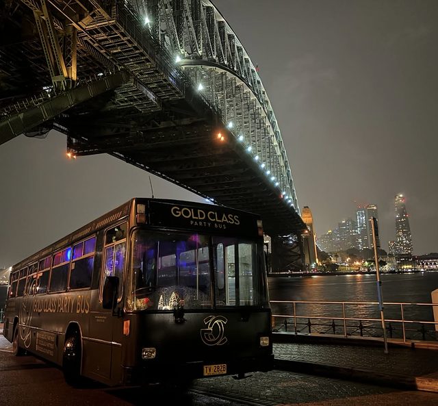 The black Gold Class party bus parked at night under the Sydney Harbour Bridge, with the city lights across the water